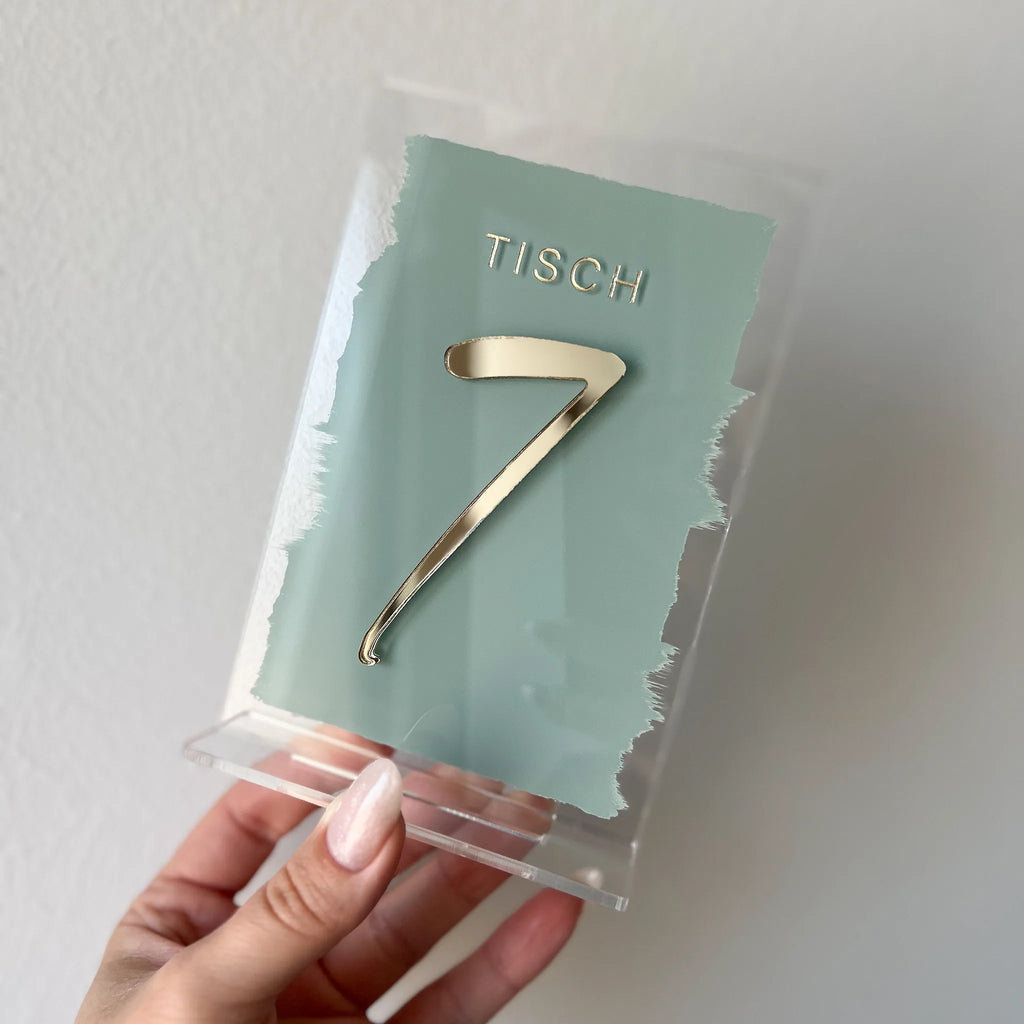 Tischnummer aus Acrylglas mit salbeigrünem Pinselstrich und goldener 3D-Zahl 7, in der Hand gehalten