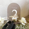 Tischnummer aus durchgefärbtem Acrylglas in Bogenform mit weißer 3D-Zahl 2, in schwarzer Handschuh-Hand gehalten, im Hintergrund weiße Rosen.