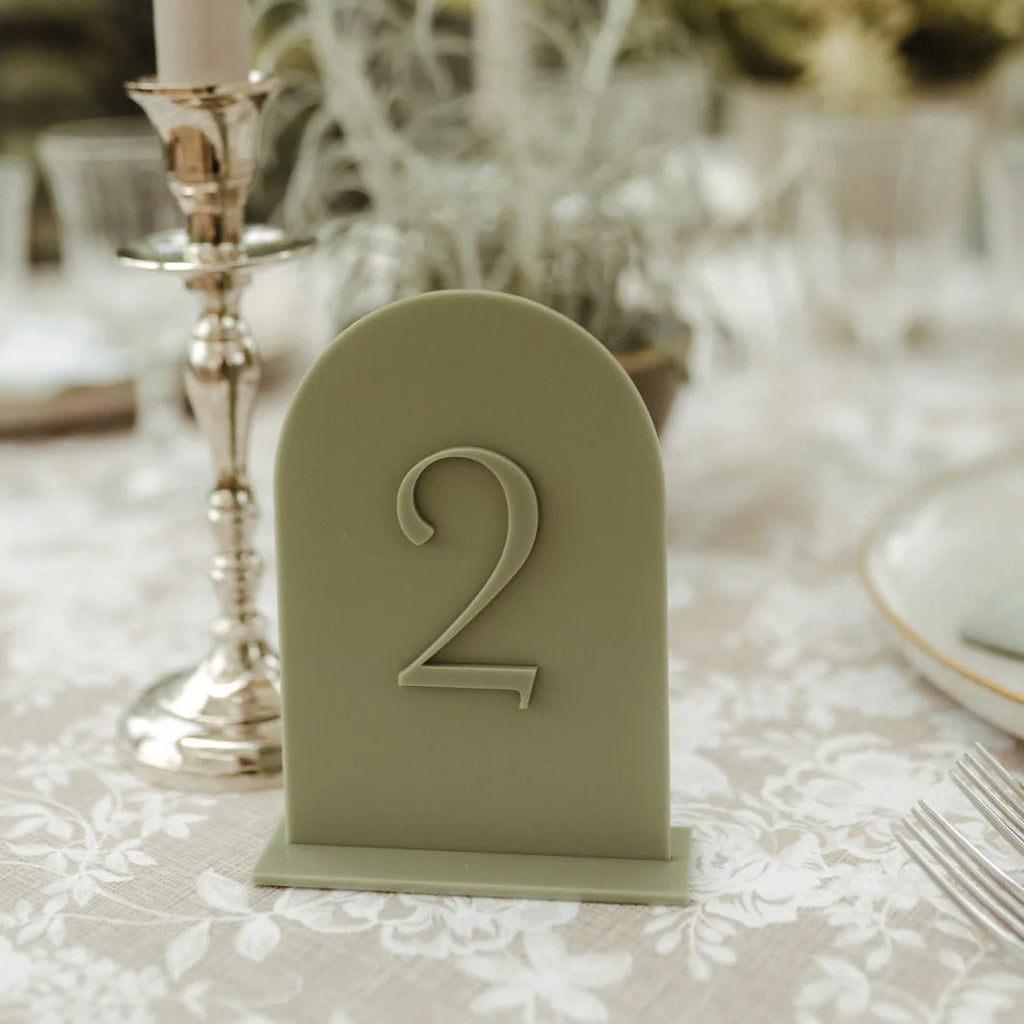 Salbeigrüne Tischnummer aus Acryl in Bogenform mit 3D-Zahl 2 auf gedecktem Hochzeitstisch neben goldenem Kerzenständer.