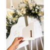 Hand hält weiße Tischnummer aus Acryl mit goldener 3D-Zahl 1 vor einem Sitzplan mit weißen Blumen im Hintergrund