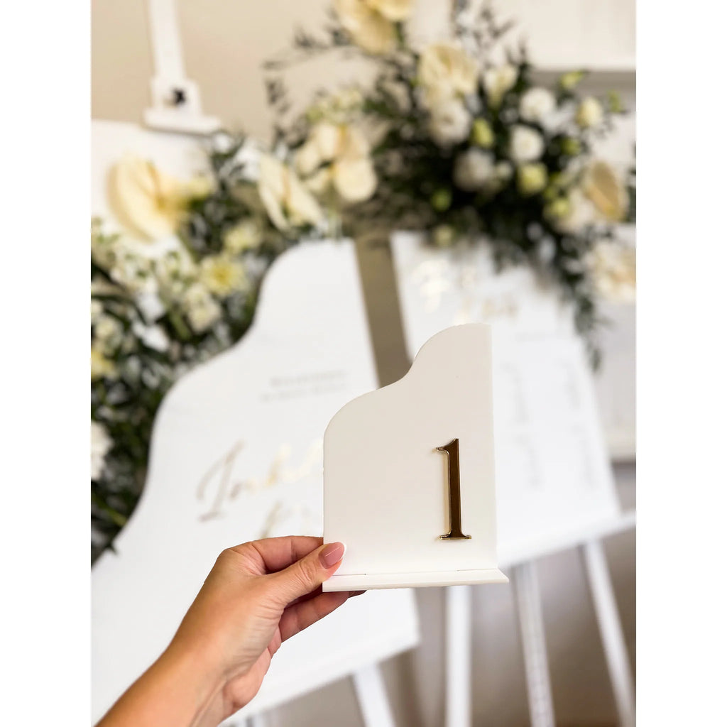 Hand hält weiße Tischnummer aus Acryl mit goldener 3D-Zahl 1 vor einem Sitzplan mit weißen Blumen im Hintergrund