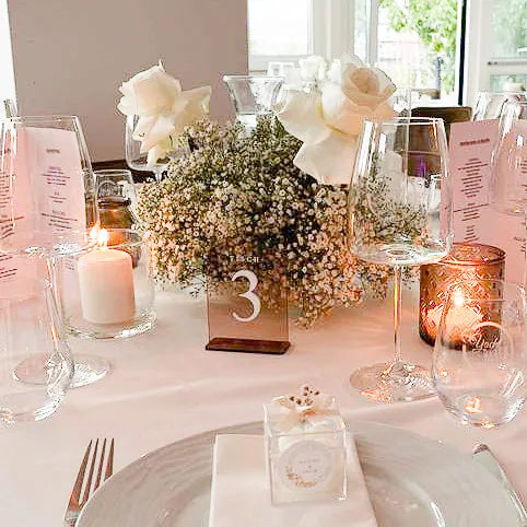 Gedeckter Hochzeitstisch mit Acrylglas-Tischnummer 3 zwischen Kerzenlicht, Schleierkraut und weißen Rosen.