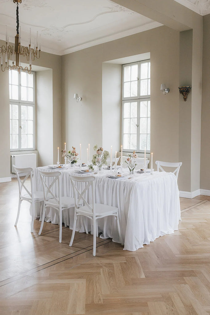 Festlich gedeckter Hochzeitstisch mit weißem Leinen, Stuhlreihen und brennenden Kerzen im hellen Saal
