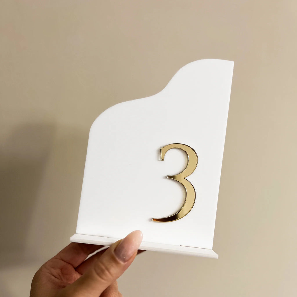 Weiße Tischnummer aus Acryl in geschwungener Form mit goldener 3D-Zahl 3, gehalten vor beigem Hintergrund