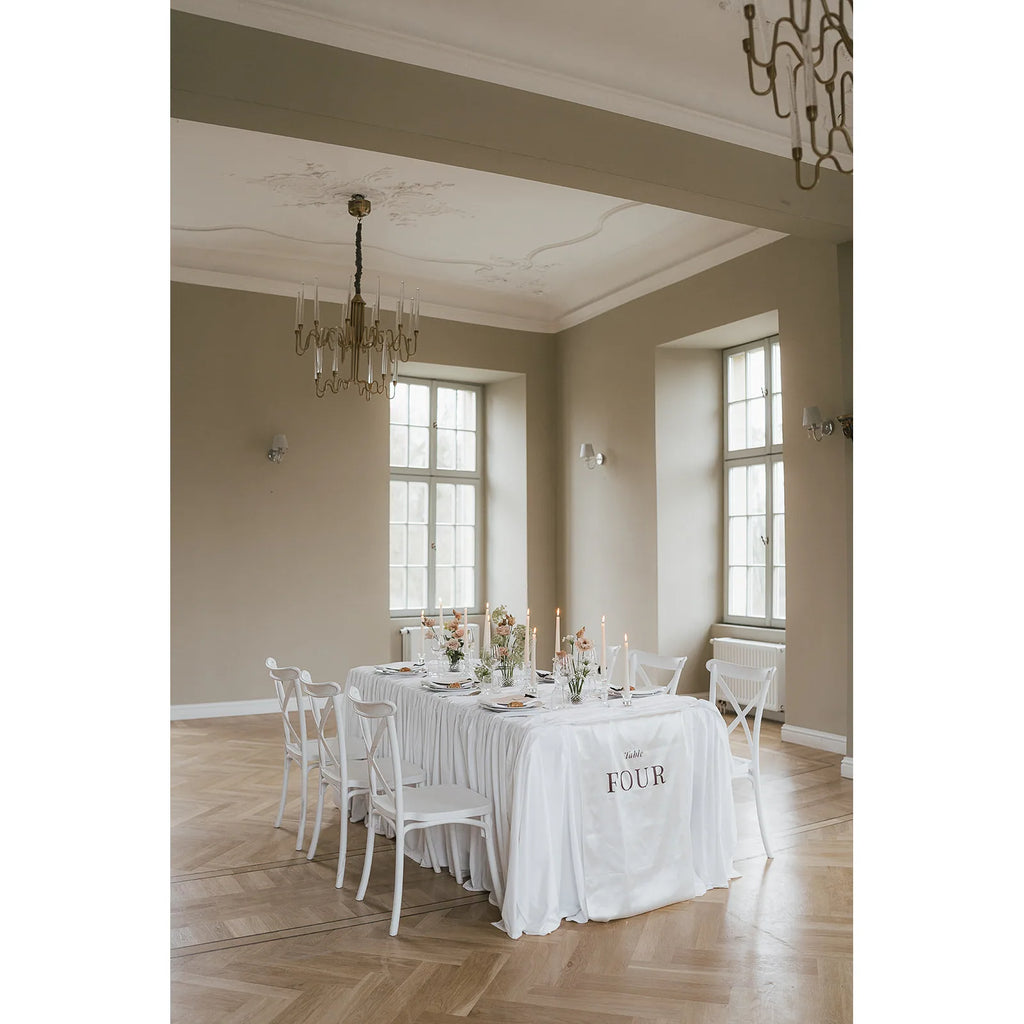 Blick in einen hellen Saal mit Stuckdecke und langem Hochzeitstisch, an dessen Kante ein Stoffbanner mit Tischnummer Four hängt