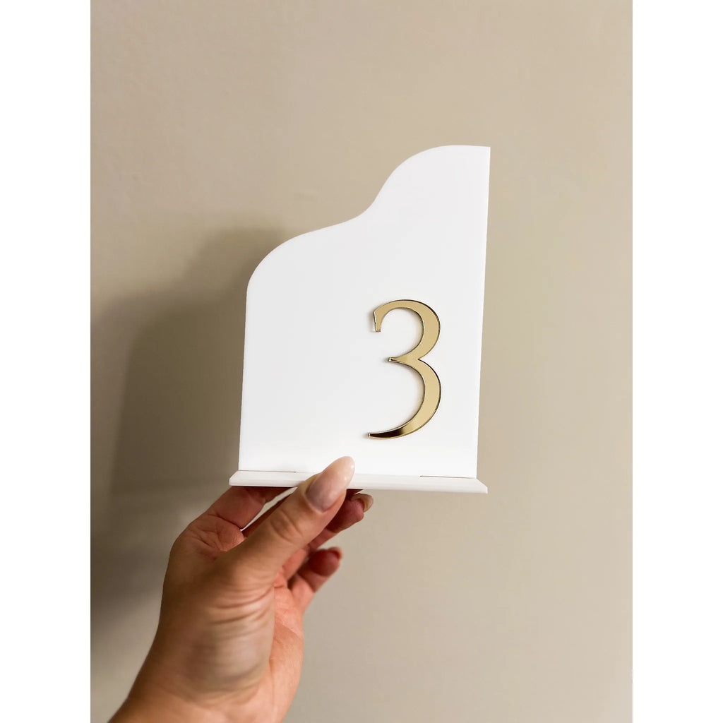 Nahaufnahme einer weißen Acryl-Tischnummer mit goldener 3D-Zahl 3 und passendem Fuß in einer Hand