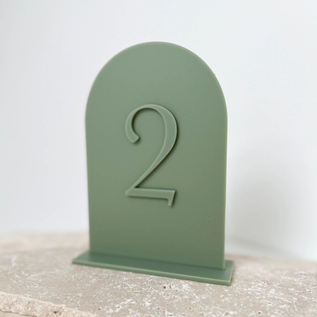 Salbeigrüne Acryl-Tischnummer mit 3D-Zahl 2 auf hellem Steinuntergrund, Nahaufnahme der Bogenform.