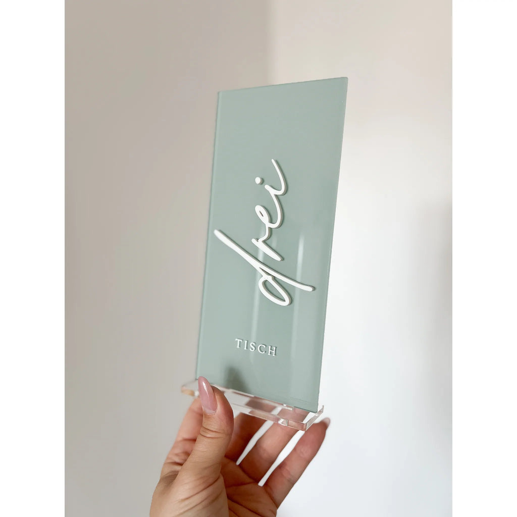 Salbeigrüne Tischnummer mit dem Schriftzug Drei in weißer 3D-Schrift und transparentem Acrylfuß, schräg von der Seite in einer Hand gehalten.
