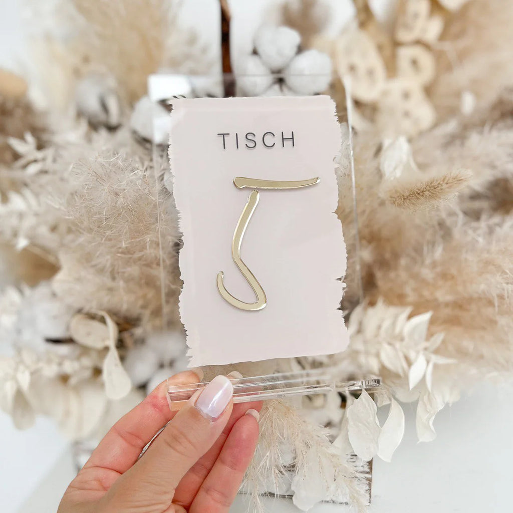 Nahaufnahme der Tischnummer mit rosa Pinselstrich und goldener 3D-Zahl, in der Hand gehalten vor Trockenblumenstrauß