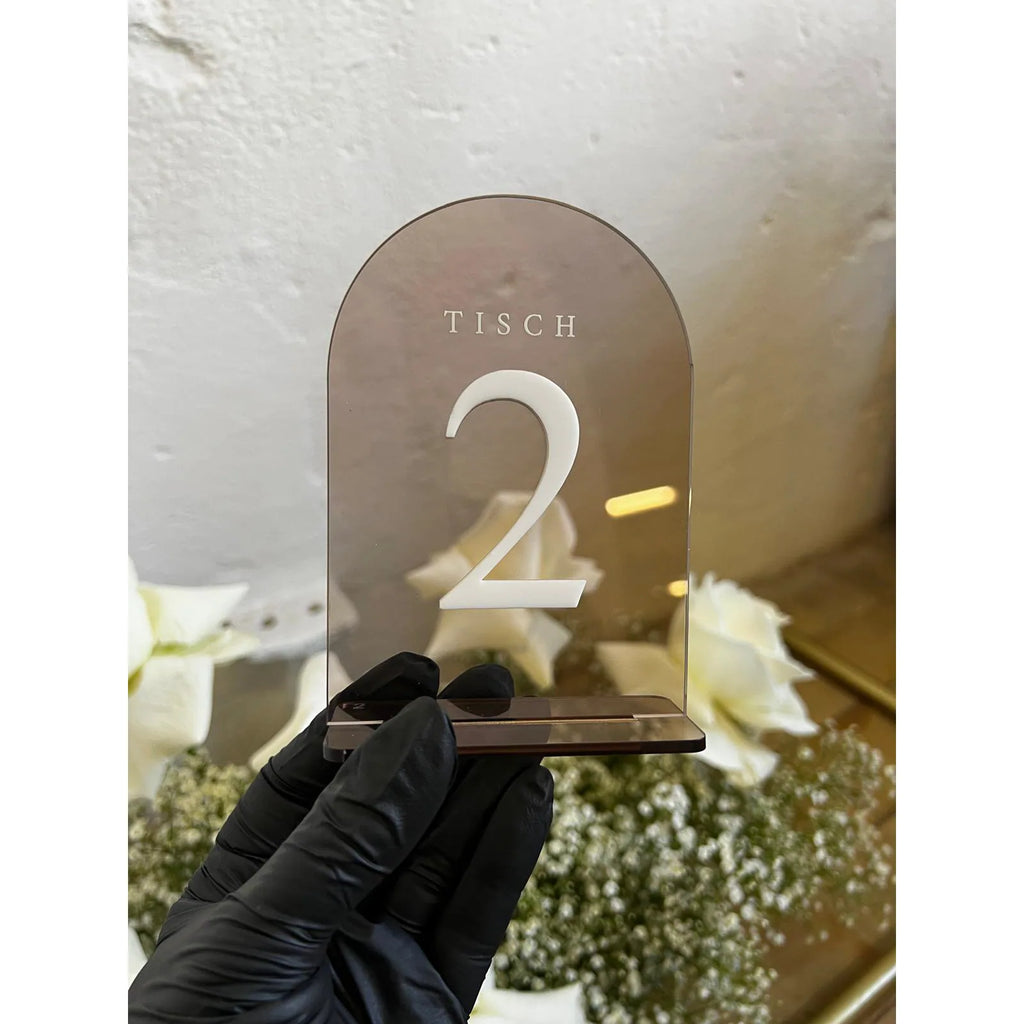 Tischnummer in Bogenform aus transparent-braunem Acrylglas mit Standfuß, freigestellt vor heller Wand.