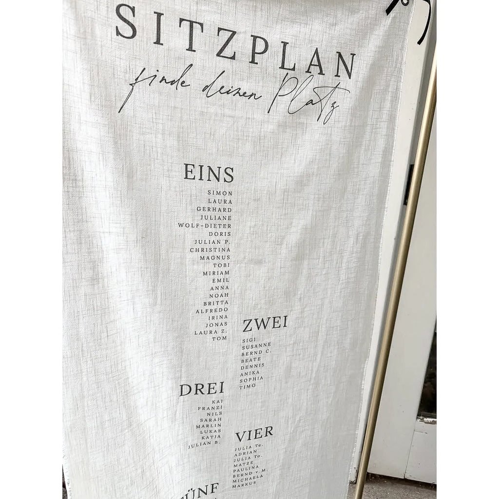 Nahaufnahme des Sitzplans Hochzeit aus Leinen mit der Überschrift Sitzplan, finde deinen Platz und den Tischnummern Eins bis Vier samt Namenslisten in schwarzer Schrift auf naturfarbenem Stoff.