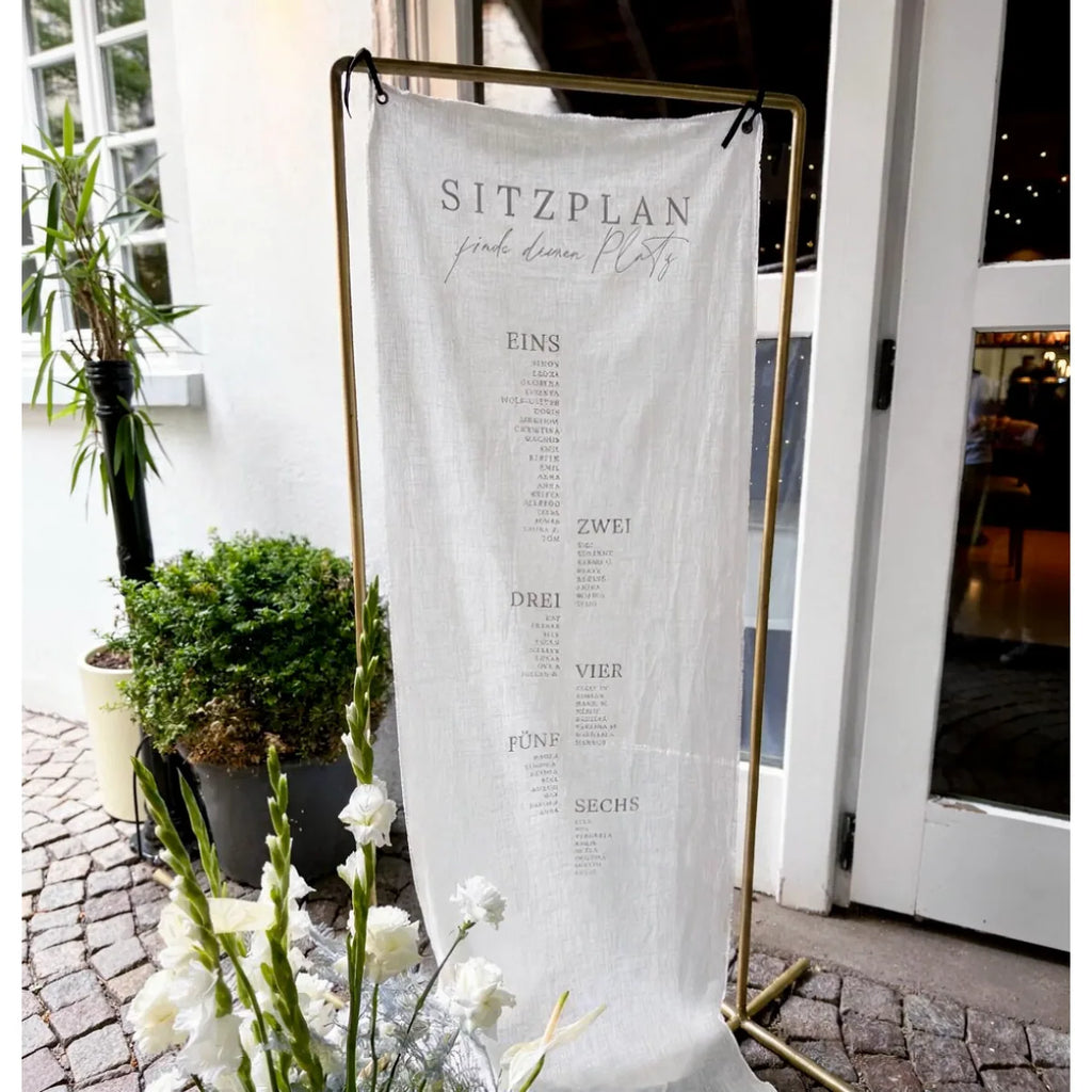 Sitzplan Hochzeit aus Leinen am goldenen Ständer vor einem Hauseingang, davor weiße Blumen und ein Kübel mit Buchsbaum.