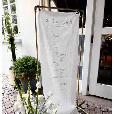 Sitzplan Hochzeit aus Leinen am goldenen Ständer vor einem Hauseingang, davor weiße Blumen und ein Kübel mit Buchsbaum.