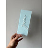 Hellblaue Tischnummer Lea aus Acrylglas mit dem Schriftzug Zwei in weißer 3D-Schrift und der Prägung Tisch, in einer Hand gehalten vor heller Wand.