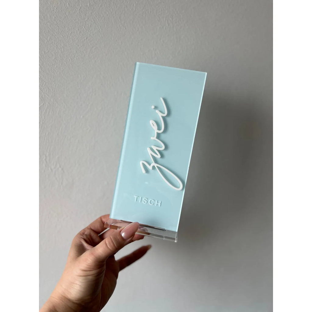 Hellblaue Tischnummer Lea aus Acrylglas mit dem Schriftzug Zwei in weißer 3D-Schrift und der Prägung Tisch, in einer Hand gehalten vor heller Wand.