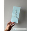Hellblaue Tischnummer Lea mit dem Schriftzug Zwei in weißer 3D-Schrift und Prägung Tisch wird schräg vor heller Wand in einer Hand gehalten, mit Farbhinweis Dusty Blue.