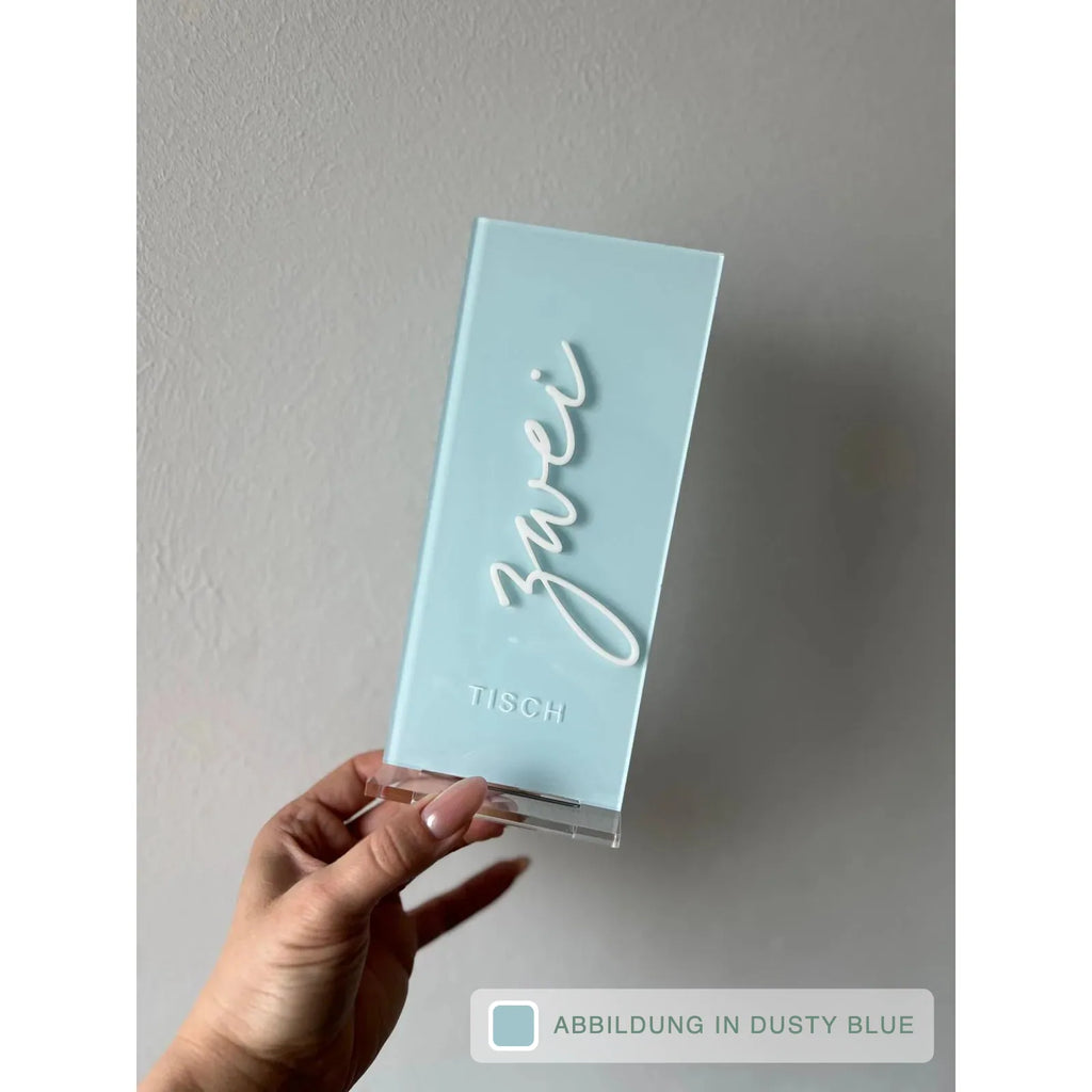 Hellblaue Tischnummer Lea mit dem Schriftzug Zwei in weißer 3D-Schrift und Prägung Tisch wird schräg vor heller Wand in einer Hand gehalten, mit Farbhinweis Dusty Blue.