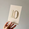 Cremefarbene Tischnummer mit spiegelgoldener 3D-Zahl 10 und Schriftzug TISCH, in der Hand gehalten