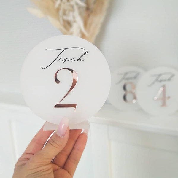 Hand hält runde Tischnummer aus satiniertem Acrylglas mit roségoldener 3D-Zahl 2 und Schriftzug Tisch.
